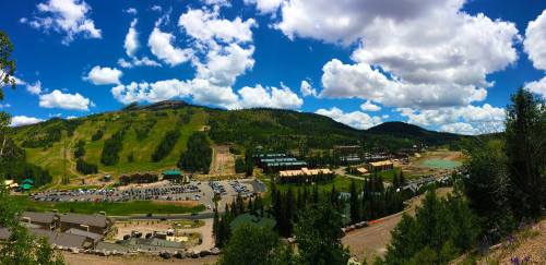 A panoramic view of a mountainous landscape with lush greenery, a blue sky, and scattered clouds, featuring a resort area below.