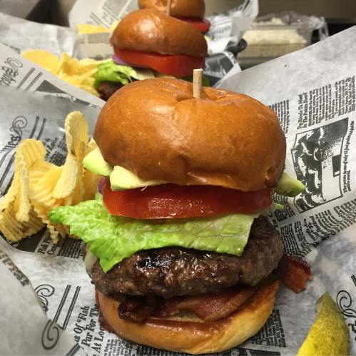 A close-up of a juicy burger with lettuce, tomato, avocado, and bacon, served with potato chips on the side.