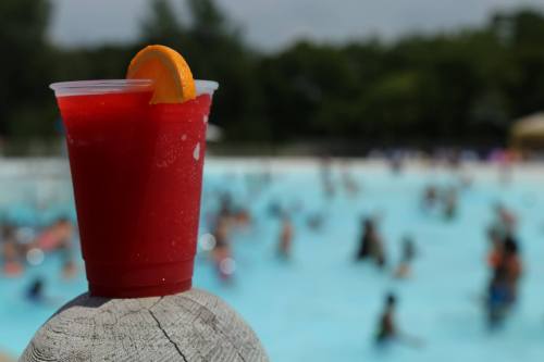 A refreshing red drink with an orange slice sits on a wooden post, with a busy pool scene in the background.