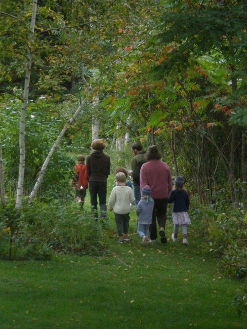 A group of children and adults walking along a wooded path surrounded by greenery and autumn foliage.
