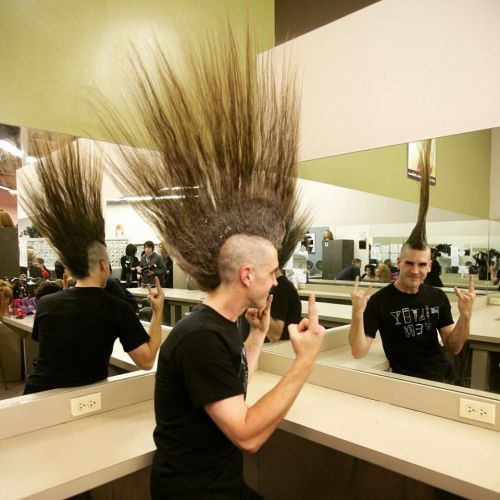A person with an exaggerated mohawk hairstyle poses in front of a mirror, making a playful gesture.