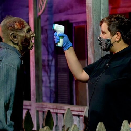 A person in a mask uses a thermometer on a zombie-like figure in a spooky setting.