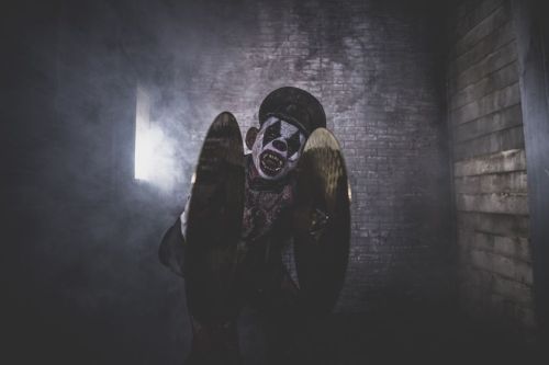 A creepy clown with a painted face crouches in a dimly lit room, holding cymbals amidst swirling smoke.