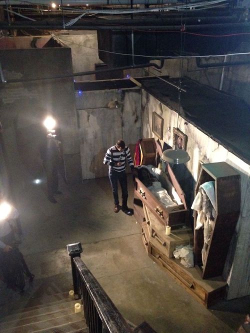 A dimly lit basement with a person standing near a coffin, surrounded by old furniture and shadows.
