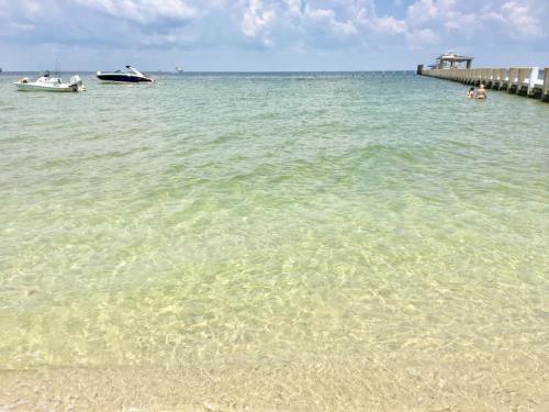 A serene beach scene with clear water, boats in the distance, and a pier under a partly cloudy sky.