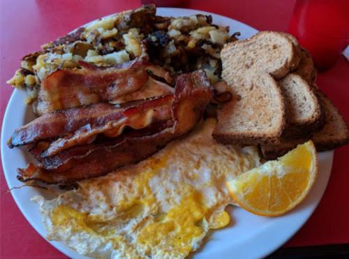 A plate of breakfast featuring crispy bacon, fried eggs, hash browns, and slices of toast with an orange wedge.