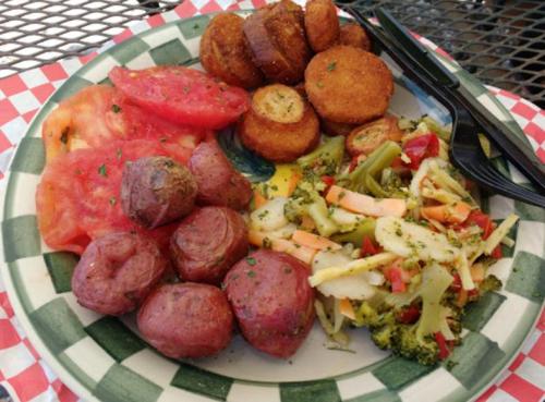A plate of food featuring sliced tomatoes, red potatoes, fried plantains, and mixed vegetables on a checkered tablecloth.