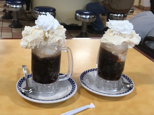 Two frosty mugs of root beer floats topped with whipped cream and scoops of ice cream, served on plates with spoons.