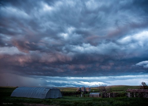 A dramatic sky filled with dark clouds over a rural landscape, featuring a barn and silos in the foreground.