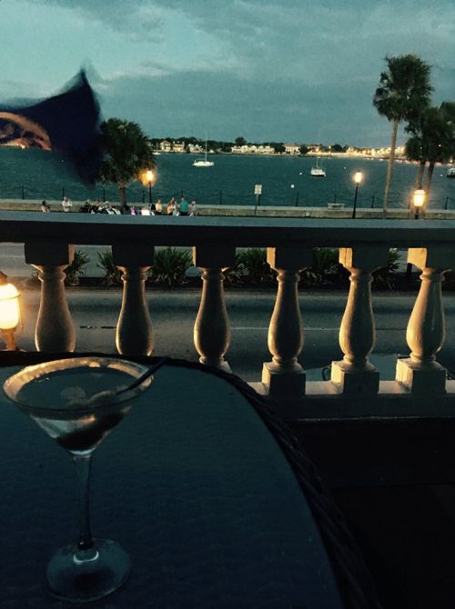 A dimly lit scene featuring a martini glass on a table, with a waterfront view and palm trees in the background.