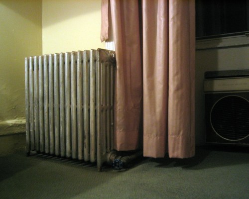 A vintage metal radiator next to a pink striped curtain in a room with carpeted flooring.