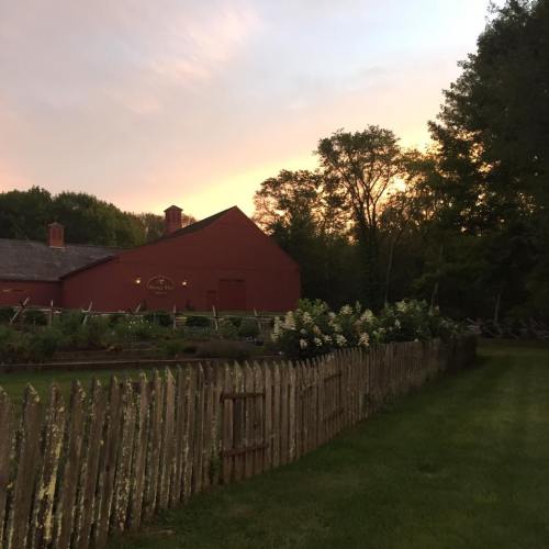A red barn at sunset, surrounded by greenery and a wooden fence, with trees in the background.