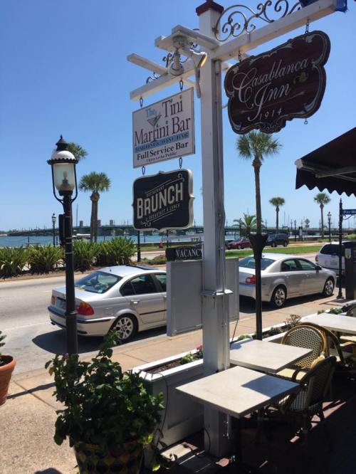 A sunny street view featuring a sign for a martini bar and a brunch spot, with palm trees and cars in the background.