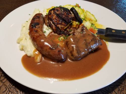 A plate of grilled sausage, beef, and scallops served with mashed potatoes and mixed vegetables, drizzled with gravy.