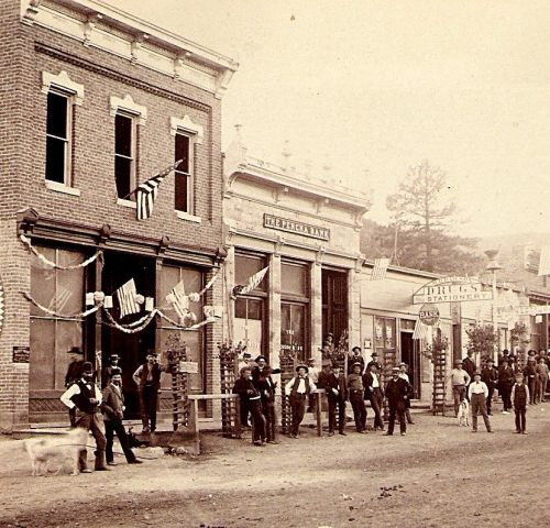 Historic street scene with people and buildings decorated with flags, showcasing a bank and shops in a small town.