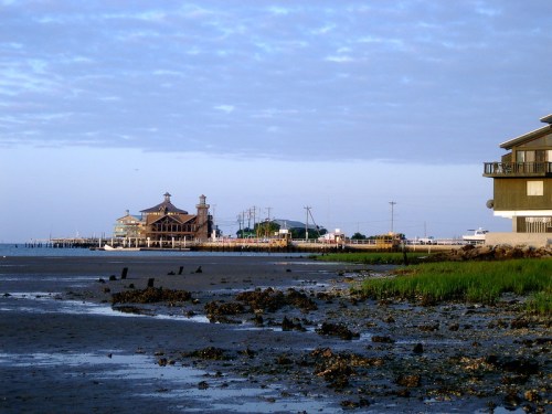 A coastal scene featuring a wooden building, sandy shore, and distant structures under a cloudy sky.