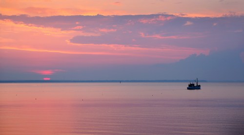 A serene sunset over calm waters, with a lone fishing boat silhouetted against vibrant pink and purple skies.