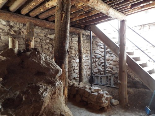 Interior of a rustic structure with wooden beams, stone walls, and a staircase leading to an upper level.