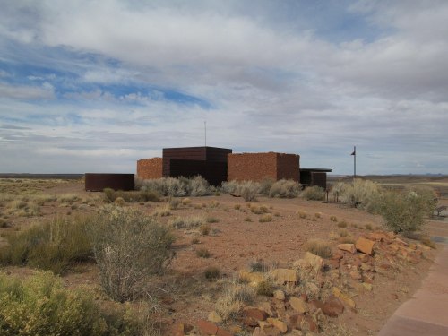 A modern building made of stone and metal, surrounded by desert vegetation under a cloudy sky.