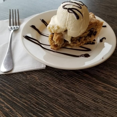 A slice of cookie pie topped with vanilla ice cream and drizzled with chocolate sauce on a white plate.
