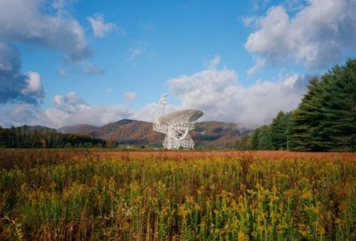 A large satellite dish stands in a colorful field, surrounded by mountains and trees under a blue sky with clouds.