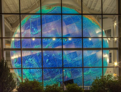 A large, illuminated globe with colorful topographic details, viewed through a glass wall at night.