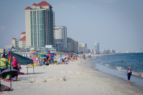 A busy beach scene with colorful umbrellas, people enjoying the sun, and tall buildings in the background.