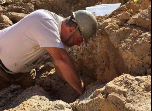 A man in a hard hat digs in a sandy excavation site, focused on his work.