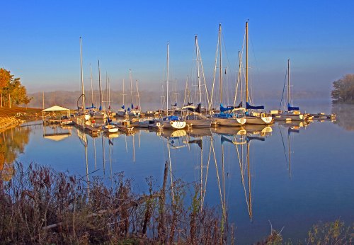 A serene marina at dawn, with sailboats reflected in calm water and mist rising in the background.
