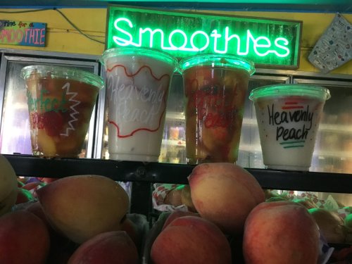 Four smoothie cups with colorful labels sit on a shelf, surrounded by fresh peaches, under a glowing "Smoothies" sign.