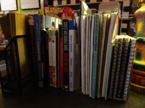 A collection of books and notebooks arranged on a shelf, with a blurred background of colorful decor.