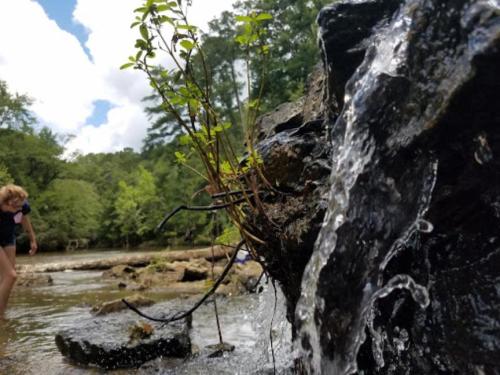 A small waterfall flows over rocks, with a child playing in the background near a stream surrounded by greenery.