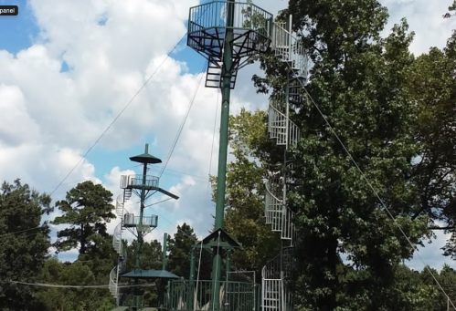 A tall green climbing structure with multiple levels and spiral staircases, surrounded by trees and a blue sky.
