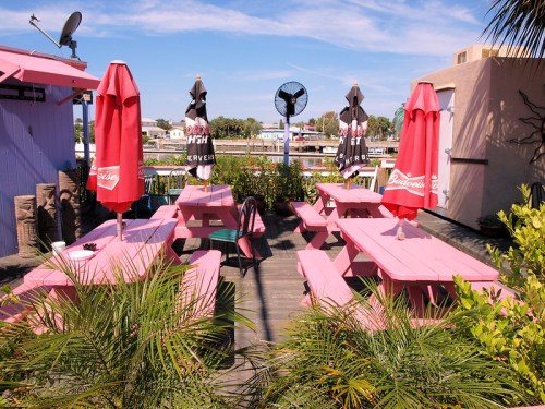 A vibrant outdoor seating area with pink picnic tables, red umbrellas, and lush greenery, overlooking a waterway.
