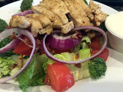 A fresh salad topped with grilled chicken, red onions, tomatoes, broccoli, and a side of dressing.