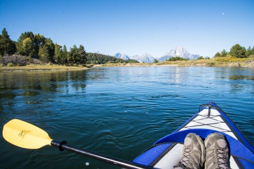 A view from a kayak on a calm river, with mountains and trees in the background under a clear blue sky.