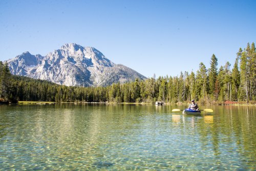 A person kayaking on a clear lake surrounded by trees and mountains under a blue sky.