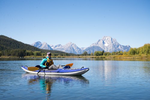 A person in a kayak on a calm lake, surrounded by mountains and clear blue skies.