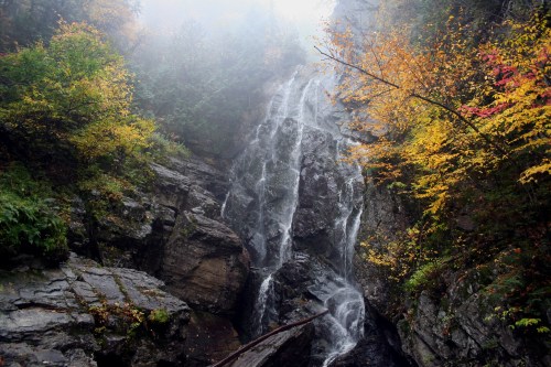 A misty waterfall cascades down rocky cliffs, surrounded by vibrant autumn foliage in a serene forest setting.