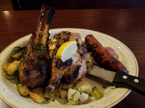 A plate of grilled meats including lamb chops, chicken, and sausage, garnished with lemon and served with vegetables.
