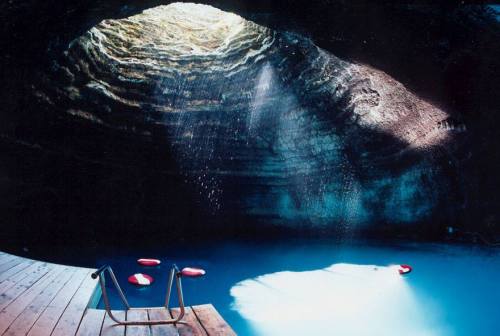 A serene underground pool with a wooden deck, illuminated by sunlight streaming through a circular opening above.