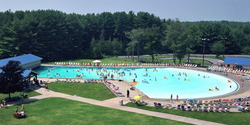 Aerial view of a large swimming pool surrounded by trees, with people swimming and lounging on deck chairs.