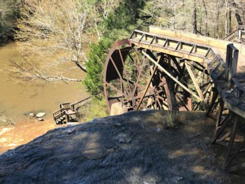 A wooden waterwheel beside a river, surrounded by trees and rocky terrain, with a wooden walkway in the background.