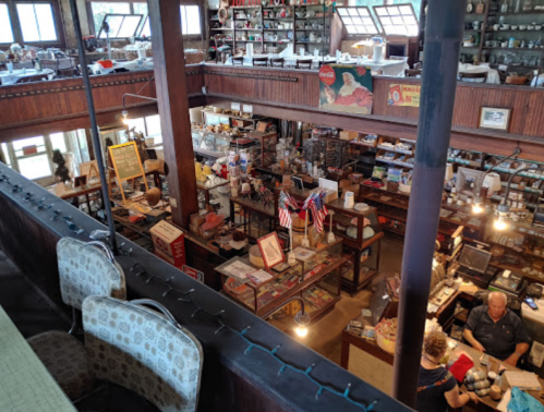 A vintage store interior filled with various items, shelves, and a couple of people at the counter.