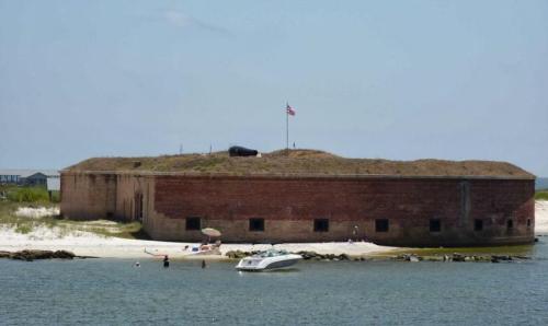 Historic brick fort on a sandy beach, with a flag and a cannon on top, surrounded by water and a boat nearby.