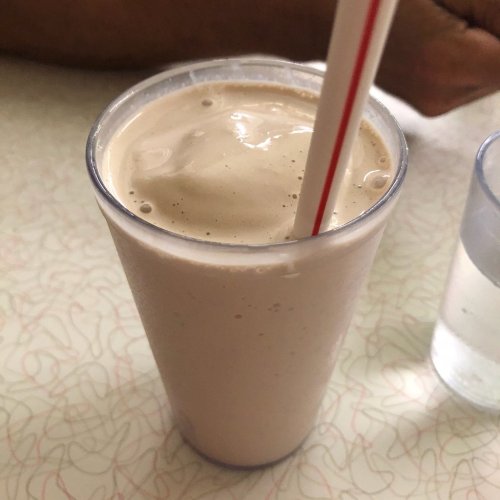 A frosty milkshake in a clear cup with a red straw, sitting on a table next to a glass of water.