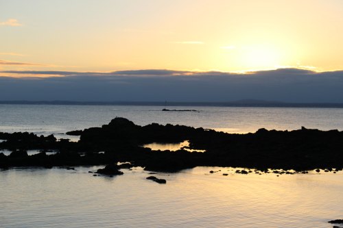 Sunset over calm waters, with rocky silhouettes and a distant lighthouse against a colorful sky.