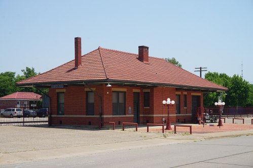 Historic red brick train station with a tiled roof, surrounded by a parking area and green trees under a clear blue sky.