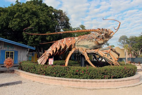 Giant lobster sculpture in a garden setting, with a blue building and trees in the background.