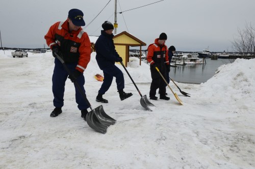 Four workers in orange and blue uniforms shovel snow near a harbor on a cloudy day. Boats are visible in the background.